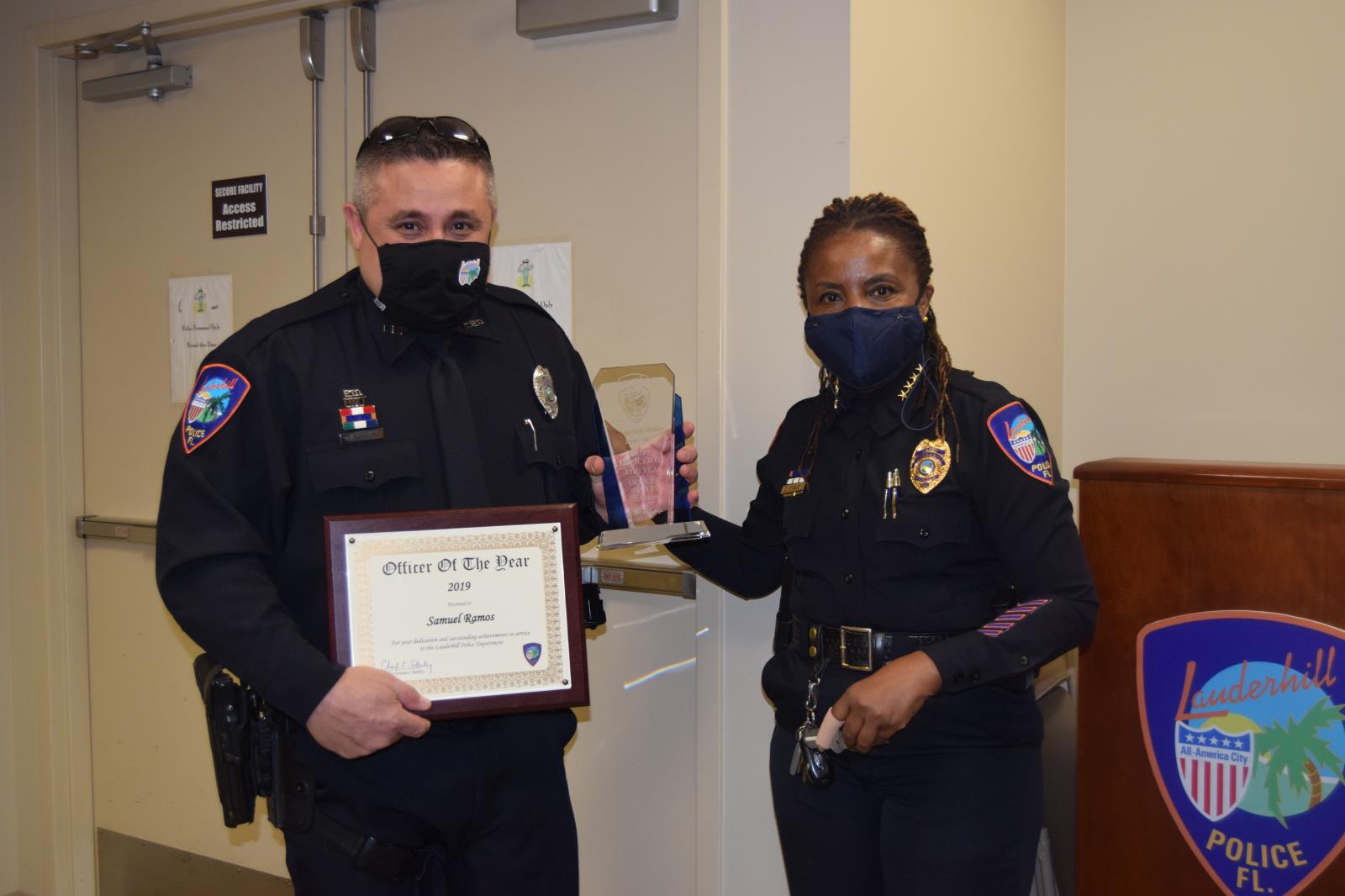 Officer Ramos and chief Stanley with certificate and award indoors beside Lauderhill police department podium