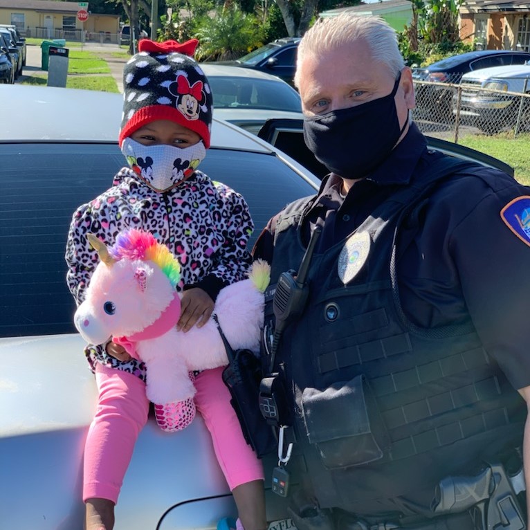 Officer yopps with young girl seated on car trunk with a plush pink unicorn toy outdoors