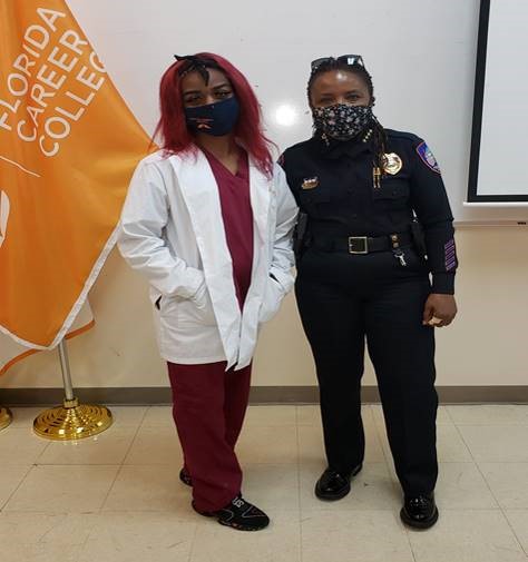 Chief Stanley with a student in a white lab coat and scrubs indoors in front of florida career college flag