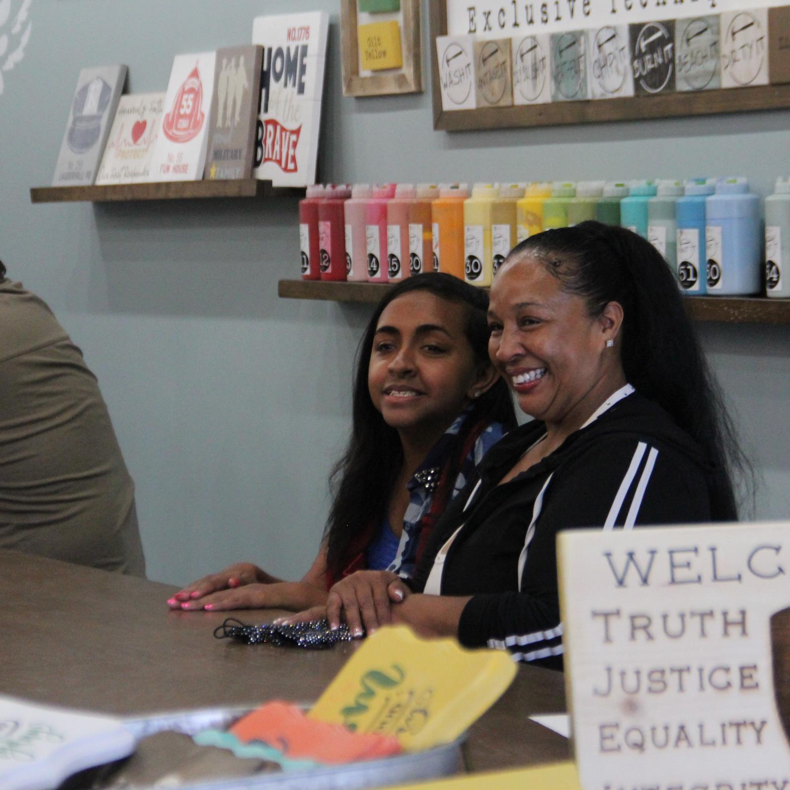 A girl and a woman smiling and sat beside each other in front of a wall with shelves of crafts