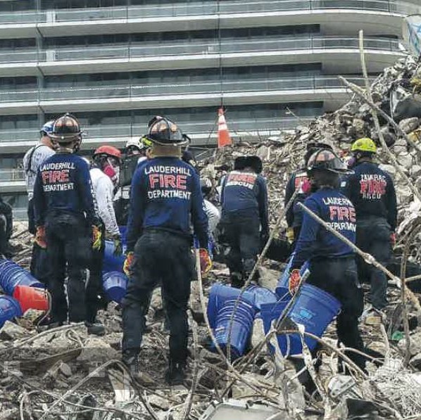 The backsides of five Lauderhill fire rescuers standing with buckets and tools among construction debris at the base of existent champlain tower