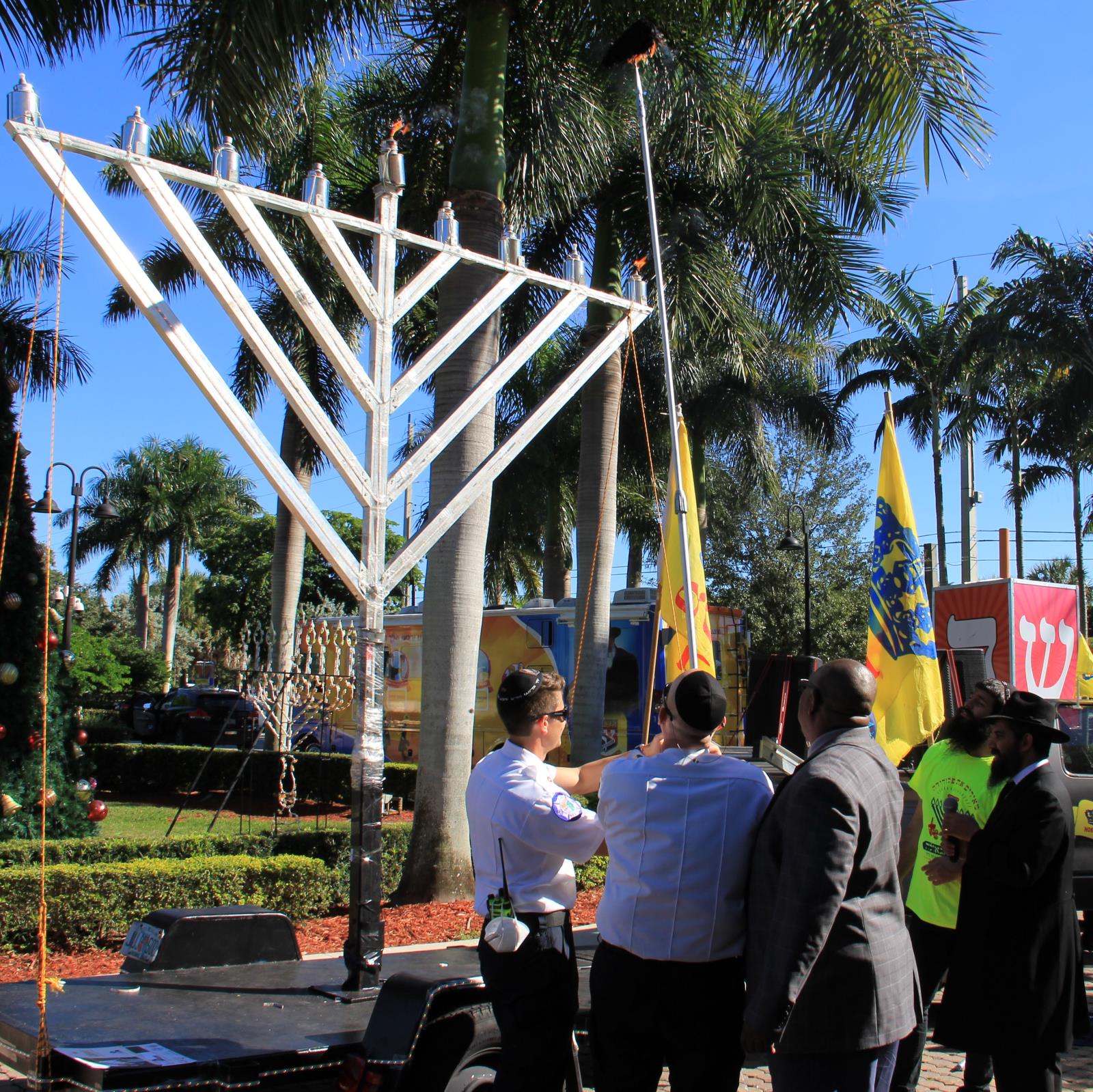 Group of people outside lighting menorah in city hall parking lot with enlarged dreidel in background