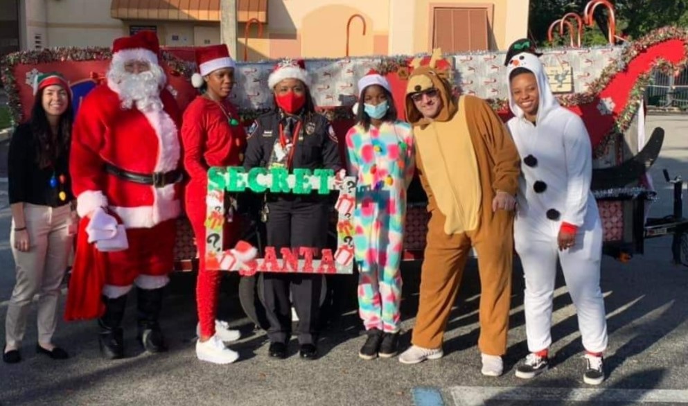 Chief Stanley standing beside 6 lauderhill officers outside in Lauderhill police department parking lot wearing holiday themed outfits in front of a decorated sleigh