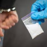 Handcuffed man and small bag of drug substance held by blue gloved hand